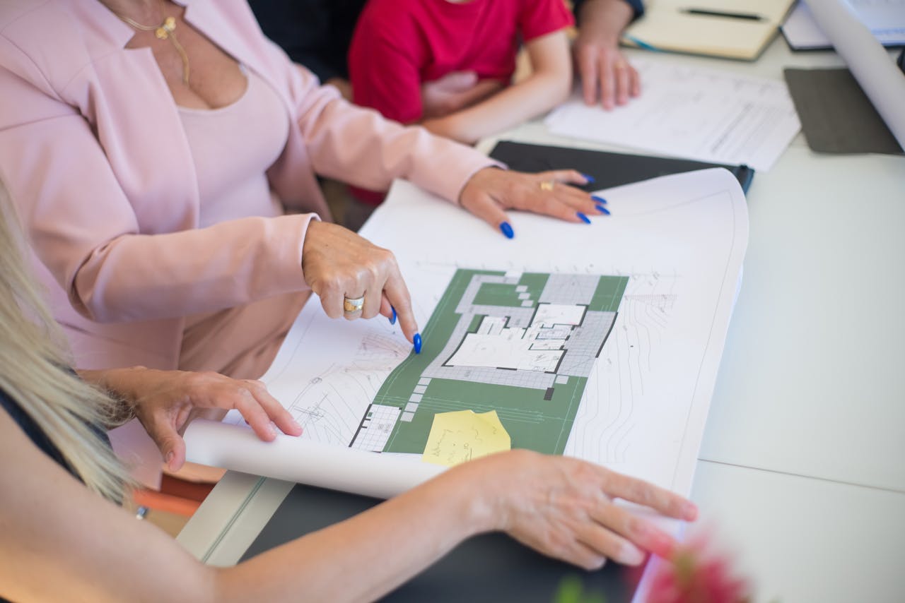 A real estate agent in a pink blazer shows floor plans to clients, discussing property details.